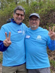 Two men in bright blue shirts posing outdoors, making peace signs with their hands, smiles on their faces amid green bushes.